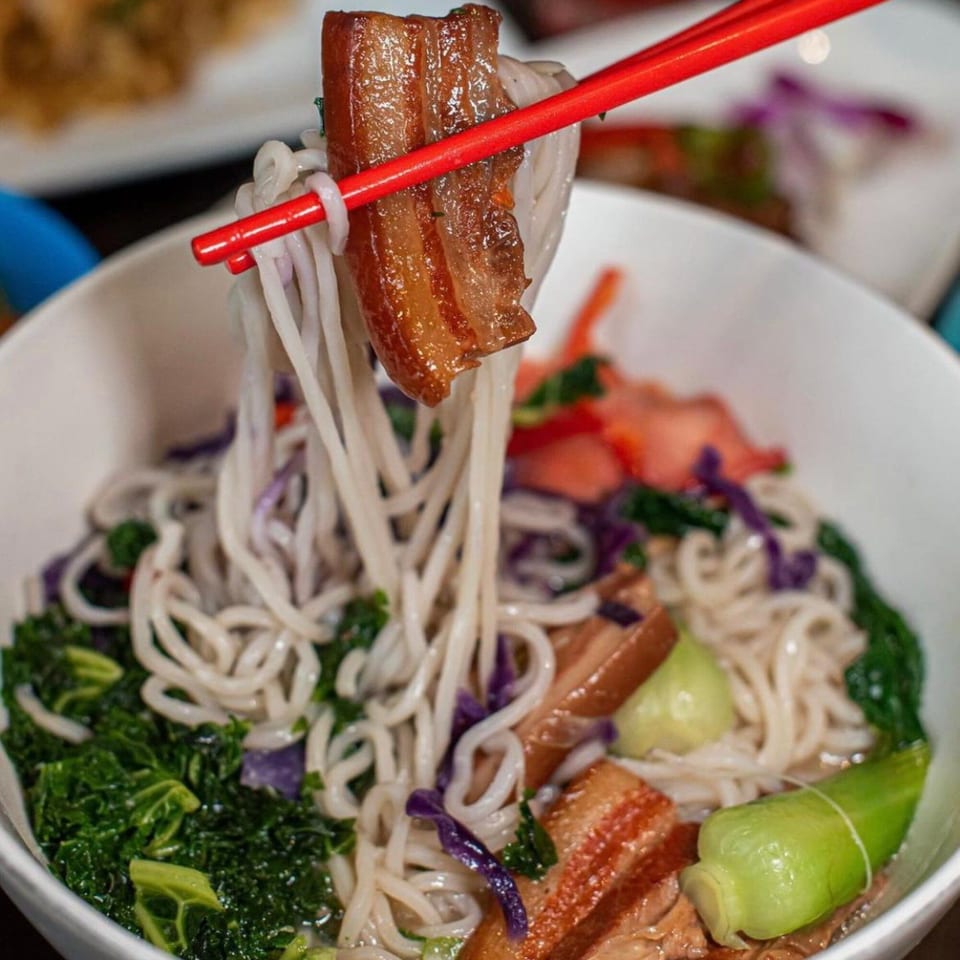 Red chopsticks lift a piece of pork belly and noodles from a bowl of Reren Ramen.