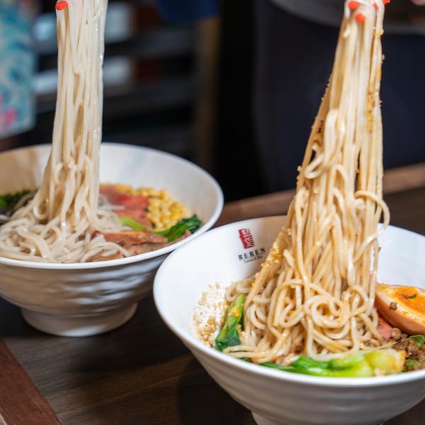 Chopsticks lifting long, thin ramen noodles from two bowls of Reren Lamen, garnished with egg and greens.