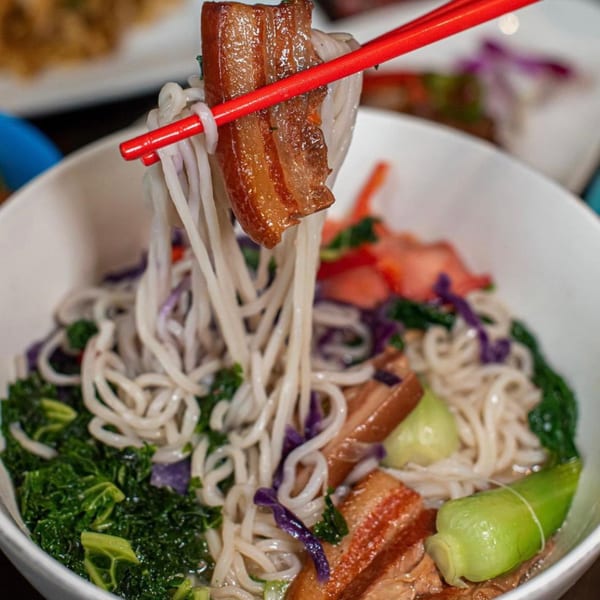 Red chopsticks lift noodles and pork belly from a bowl of Reren Lamen with vegetables.