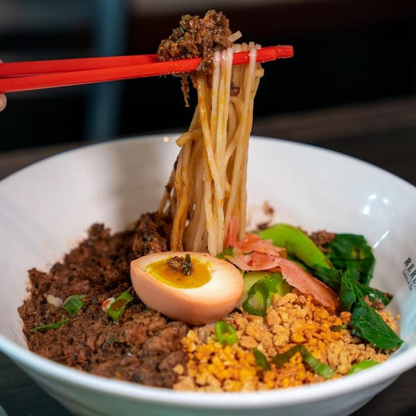 Chopsticks lifting ramen noodles with minced meat, a soft-boiled egg, and greens from a white bowl.