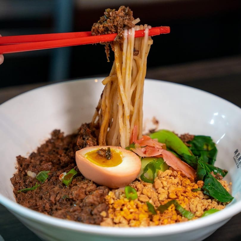 Chopsticks lifting ramen noodles with minced meat, a soft-boiled egg, and greens from a white bowl.