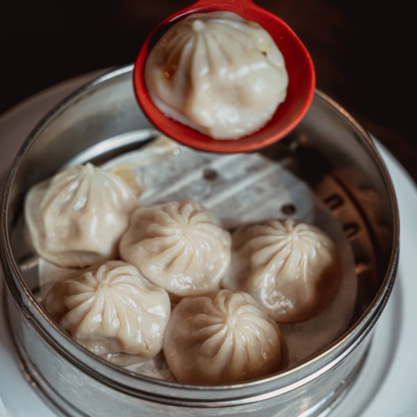 A red spoon lifts a soup dumpling from a steamer basket filled with several more.